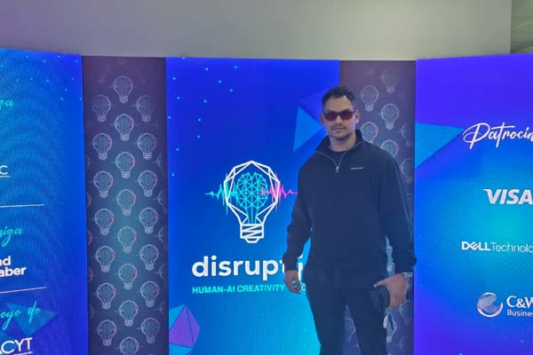 Chiragx frente al photocall de Disrupt IT 2026 (Human-AI Creativity) en Ciudad del Saber, con chaqueta de CryptoManji y lentes rojos